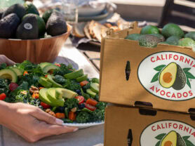 Close-up of hands serving a California Avocado superfood salad on a farm table next to cardboard boxes filled with California Avocados