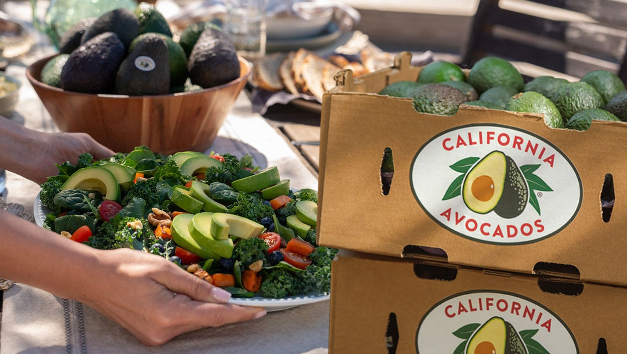 Close-up of hands serving a California Avocado superfood salad on a farm table next to cardboard boxes filled with California Avocados